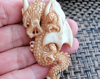 May include: A detailed, cream-colored dragon carving with intricate scales, horns, and wings. The dragon is carved from a light-colored material, possibly bone or ivory, and features a curled tail and detailed facial features. The carving is a decorative item.