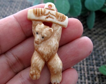 May include: A carved pendant featuring a bear holding a log. The bear is light brown with detailed fur and small black eyes. The log is a lighter shade of brown with carved details. The pendant has a small hole at the top for a chain.
