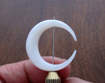 May include: A white crescent moon shaped piece of jewelry is displayed on a stand. The jewelry is made of a smooth, white material and has a hole in the center for a pin or earring post.