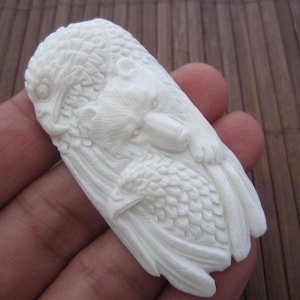 May include: White carved bone pendant featuring a bear and two birds. The bear is in the center of the pendant and the birds are on either side of the bear. The pendant is intricately carved with details on the bear's fur, the birds' feathers, and the background.