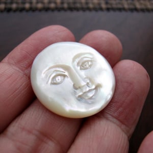 May include: A carved, oval-shaped, white mother-of-pearl pendant featuring a smiling face with detailed eyes, nose, and mouth. The iridescent surface reflects light, creating a shimmering effect. The pendant is held in a hand.