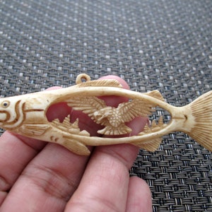 May include: A carved bone pendant in the shape of a fish with a bird flying inside. The fish is detailed with scales and fins, and the bird has spread wings.