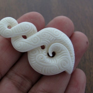 Könnte beinhalten: Cremefarbener Anhänger mit einem wirbelnden, geschnitzten Design. Der Anhänger weist komplizierte Muster und eine glatte, polierte Oberfläche auf. Das Design erinnert an eine stilisierte Welle oder Spirale mit einem Loch in der Mitte. Dieses einzigartige Stück ist wahrscheinlich handgefertigt.