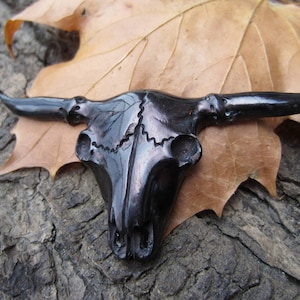 May include: A black ceramic bull skull with horns, resting on a brown leaf. The skull is highly detailed and has a glossy finish.