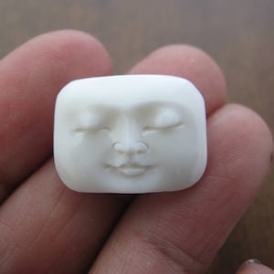 Könnte beinhalten: Eine weiße, geschnitzte Steinskulptur eines Gesichts mit geschlossenen Augen und einem kleinen Lächeln.