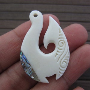 May include: White bone pendant carved in the shape of a fish hook with a mother of pearl inlay. The hook is decorated with a traditional Maori design.