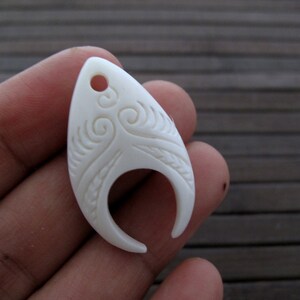 Puede incluir: Pendiente de hueso blanco con un diseño tallado. El pendiente tiene forma de lágrima con un agujero en la parte superior para colgar.