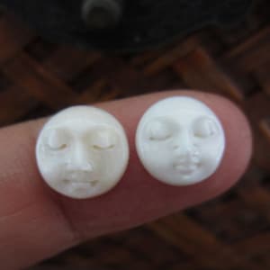 May include: Two small, round, off-white face carvings with closed eyes and serene expressions. The carvings are detailed with delicate features, suggesting a handcrafted nature. The faces are similar in design, with one appearing slightly darker than the other.