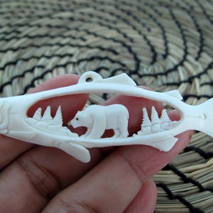 May include: A white bone carving of a fish with a polar bear carved inside. The fish is carved in a detailed style with scales and fins. The polar bear is walking through a forest scene.