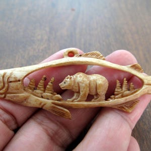 Puede incluir: Un colgante de hueso tallado en forma de pez con un oso caminando dentro del cuerpo del pez. El oso y el pez están tallados con detalle y el colgante tiene un agujero en la parte superior para colgar.