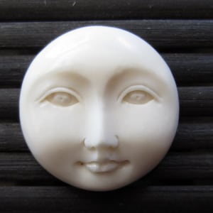 May include: A round, off-white face with delicate features, including closed eyes, a small nose, and a gentle smile. The face is smooth and appears to be made of a solid material, possibly ceramic or resin. The overall design evokes a sense of serenity.