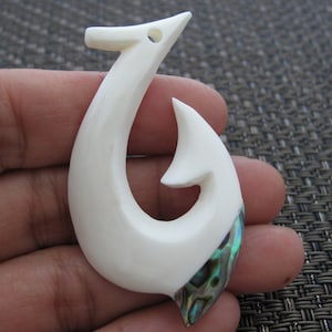 Puede incluir: Colgante de hueso blanco tallado en forma de anzuelo. Presenta una incrustación iridiscente de concha de abulón. El anzuelo tiene un diseño curvo con un pequeño agujero en la parte superior. Este colgante artesanal es una pieza de joyería única. Mide unos 8 cm de largo.