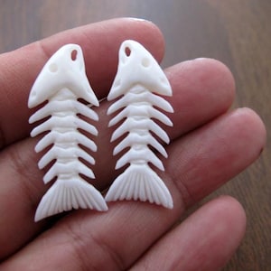 May include: Two white bone fish skeleton charms, each with a detailed carving of the bones and a tail fin.
