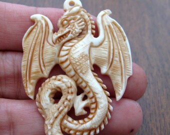 May include: Ivory-colored dragon pendant with detailed carving. The dragon has outstretched wings, a serpentine body, and a loop at the top for hanging. The pendant is likely made of bone or a similar material.