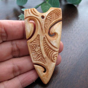 May include: A carved pendant in a shield shape, with detailed patterns and a small hole at the top. The pendant is a light tan colour, with darker brown accents in the carved details. The design includes swirling lines and geometric shapes.