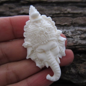 May include: White carved elephant head pendant with intricate details. The elephant is wearing a crown and has a long trunk.