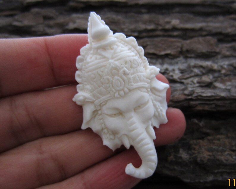 May include: White carved elephant head pendant with intricate details. The elephant is wearing a crown and has a long trunk.