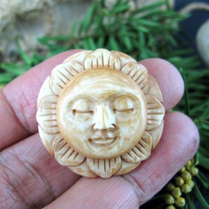 May include: A carved sun face with closed eyes and a smiling expression. The sun is surrounded by a floral design. The carving is a light beige colour and appears to be made of wood or a similar material.