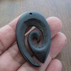May include: A dark brown, teardrop-shaped pendant with a spiral design carved into it. The pendant has a small hole at the top for a necklace cord. The wood grain is visible, and the pendant is smooth.