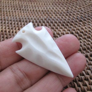 Könnte beinhalten: Elfenbeinfarbener Pfeilspitzenanhänger mit spitzer Spitze und geschnitzten Details. Der Anhänger hat ein Loch zum Auffädeln und eine strukturierte Oberfläche. Die Pfeilspitze wird in einer Hand gehalten.