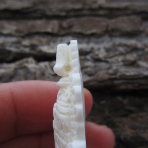May include: White carved bone pendant with intricate details. The pendant is shaped like a figure with a pointed top and a rounded bottom.