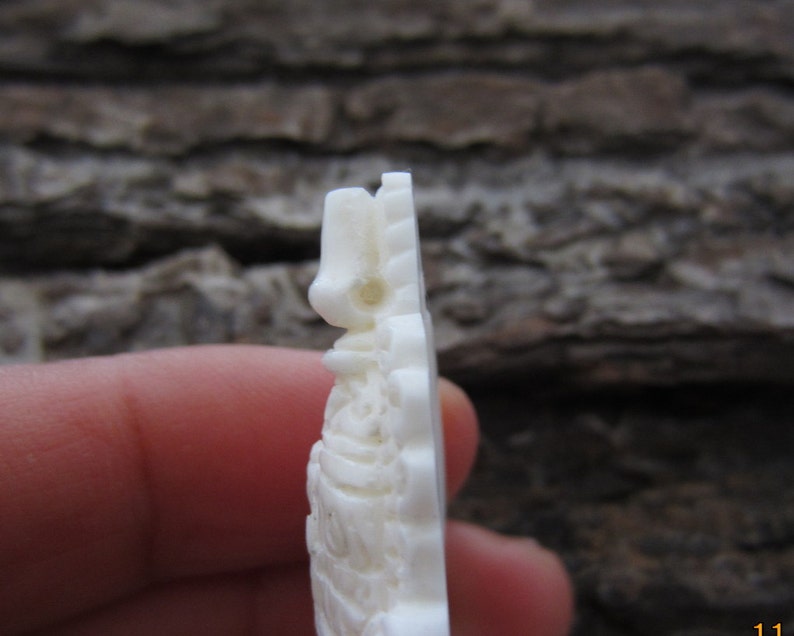 May include: White carved bone pendant with intricate details. The pendant is shaped like a figure with a pointed top and a rounded bottom.