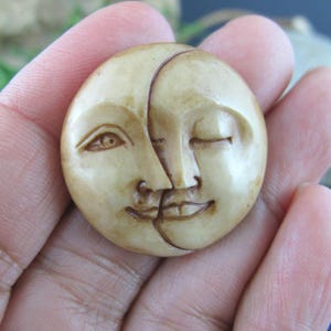 May include: A carved, circular pendant featuring two moon faces, one with an open eye and the other with a closed eye. The pendant is a light beige color and has detailed facial features.