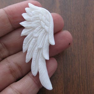May include: White carved bone angel wing pendant. The wing is detailed with feathers and has a smooth, polished finish.