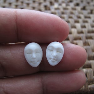 May include: Two small, white, oval-shaped face carvings with closed eyes and subtle facial features. The carvings are held in a hand, showcasing the intricate details of the miniature sculptures. The background is a woven texture.