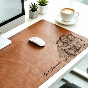 May include: A rectangular brown leather desk mat featuring a laser-engraved illustration of a parent and two children embracing. The text "Dad, I love you." is engraved in the bottom right corner. The mat is placed on a white desk surface.