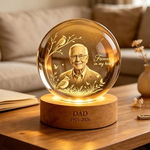 Peut inclure: Un ornement en verre sphérique présentant le portrait gravé d'une personne âgée, deux oiseaux sur des branches et le texte "Forever in my heart". La sphère repose sur une base cylindrique en bois gravée "DAD" et "1955-2026".