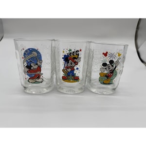 Può includere: Tre bicchieri trasparenti con grafiche colorate di Topolino. Il primo bicchiere mostra Topolino come stregone, il secondo come regista cinematografico con un ciak, e il terzo mostra Topolino che saluta con fuochi d'artificio sullo sfondo.