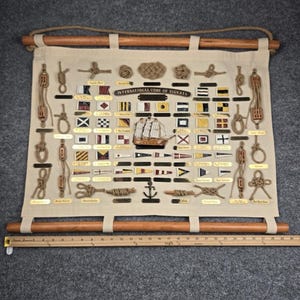 May include: A wall hanging featuring the International Code of Signals with various nautical flags, knots, and a central ship model on beige fabric. It is suspended by two wooden rods and rope. A wooden ruler shows a width of approximately 26 inches.