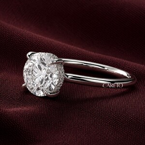 May include: A silver-toned ring featuring a large, round, faceted clear stone held by four prongs. The band is smooth and polished, with the word CARETO engraved on the side. The setting includes a small halo of tiny stones beneath the main gem.