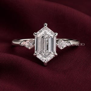 May include: A silver-toned ring featuring a large, elongated hexagonal-cut center stone held by six prongs, flanked by two smaller kite-shaped side stones. The band is smooth and polished. The text CARETO is visible on the right side of the ring.