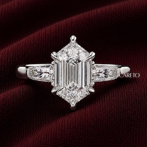 May include: A silver-toned ring featuring a large, elongated hexagonal-cut center stone held by six prongs, flanked by two smaller trapezoidal-cut side stones. The metal band is smooth and polished, with the text CARETO visible on the right side.