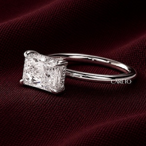 May include: A silver-toned ring featuring a large, rectangular radiant-cut clear gemstone held by four prongs. The band is smooth and polished. The word CARETO is printed in small white letters on the lower right side of the ring setting.
