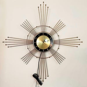 May include: A gold-toned wall clock with a black face and white numerals. The clock has a unique design with thin metal rods radiating outwards from the centre.