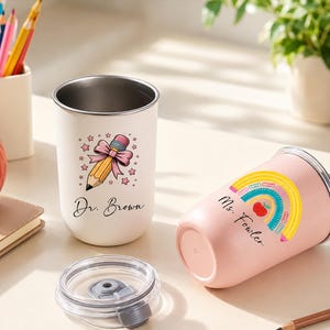 Könnte beinhalten: Ein weißer Becher mit Bleistift- und Schleifenmotiv sowie dem Text Dr. Brown und ein rosa Becher mit Regenbogen-, Bleistift- und Apfelmotiv sowie dem Text Ms. Fowler, beide mit transparenten Deckeln und Rändern aus Edelstahl versehen.