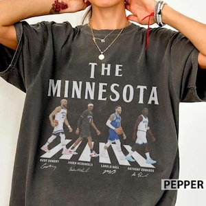 May include: A charcoal gray short-sleeve t-shirt featuring the text THE MINNESOTA and a graphic of four basketball players. Below each player are the names Rudy Gobert, Jaden McDaniels, LaMelo Ball, and a fourth player, with their signatures.