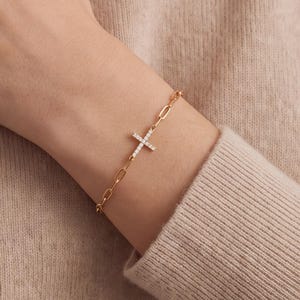 May include: A gold-toned chain link bracelet featuring a central cross charm encrusted with small, clear, sparkling stones. The bracelet is constructed from elongated oval links and secures with a clasp, resting against a beige knit fabric.
