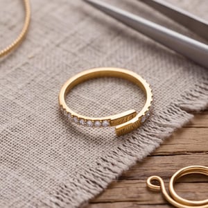 May include: A gold-toned open ring featuring a row of small, clear, round-cut stones along the band. Nearby, a pair of silver-toned metal tweezers, a thin gold-toned wire hoop, and a small gold-toned coiled wire component are visible.