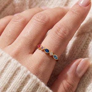May include: A gold ring featuring a series of multi-colored gemstones in various shapes, including a round red stone, a small round purple stone, a marquise-cut dark blue stone, a square white stone, and a marquise-cut light blue stone.