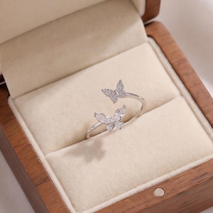 Puede incluir: Un anillo abierto de tono plateado con dos motivos de mariposa adornados con pequeñas piedras transparentes y brillantes. El anillo se muestra dentro de una caja de madera con un forro interior de terciopelo color crema, resaltando el metal y las piedras.