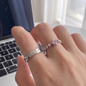 May include: Two silver-toned rings worn on fingers. One ring features a flat band with engraved star shapes and small embedded clear stones. The second ring has a central pink star-shaped stone flanked by smaller clear stones along the band.