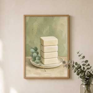 May include: A framed wall art print featuring a stack of four rectangular off-white soap bars resting on a circular ceramic dish. A single sprig of green eucalyptus leaves lies beside the soap. The background is a soft sage green watercolor wash.