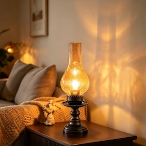 May include: A table lamp featuring a dark bronze-toned metal base and a tall, amber-colored glass shade with a distinct crackle texture. The lamp emits a warm, glowing light, casting a soft illumination and a prominent shadow on the wall behind it.