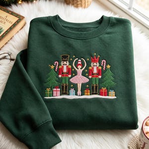 Peut inclure: Un sweat-shirt à col rond vert foncé avec une broderie représentant deux casse-noisettes entourant une ballerine en tutu rose, accompagnés de deux sapins de Noël verts, de sucres d'orge et de cadeaux colorés sur une base enneigée blanche.