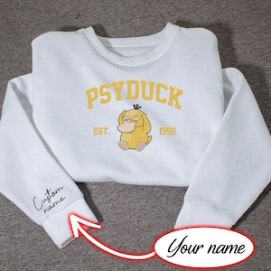 Pode incluir: Uma camisola branca de gola redonda com uma personagem Psyduck amarela bordada ao centro e o texto PSYDUCK EST. 1996 em letras amarelas. O punho da manga esquerda inclui o texto bordado a preto Custom name.