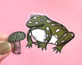 Puede incluir: Una pegatina caprichosa con una rana verde con lunares blancos y una seta verde con lunares blancos. La rana tiene una barriga blanca y está junto a la seta. La pegatina está sobre un fondo rosa.